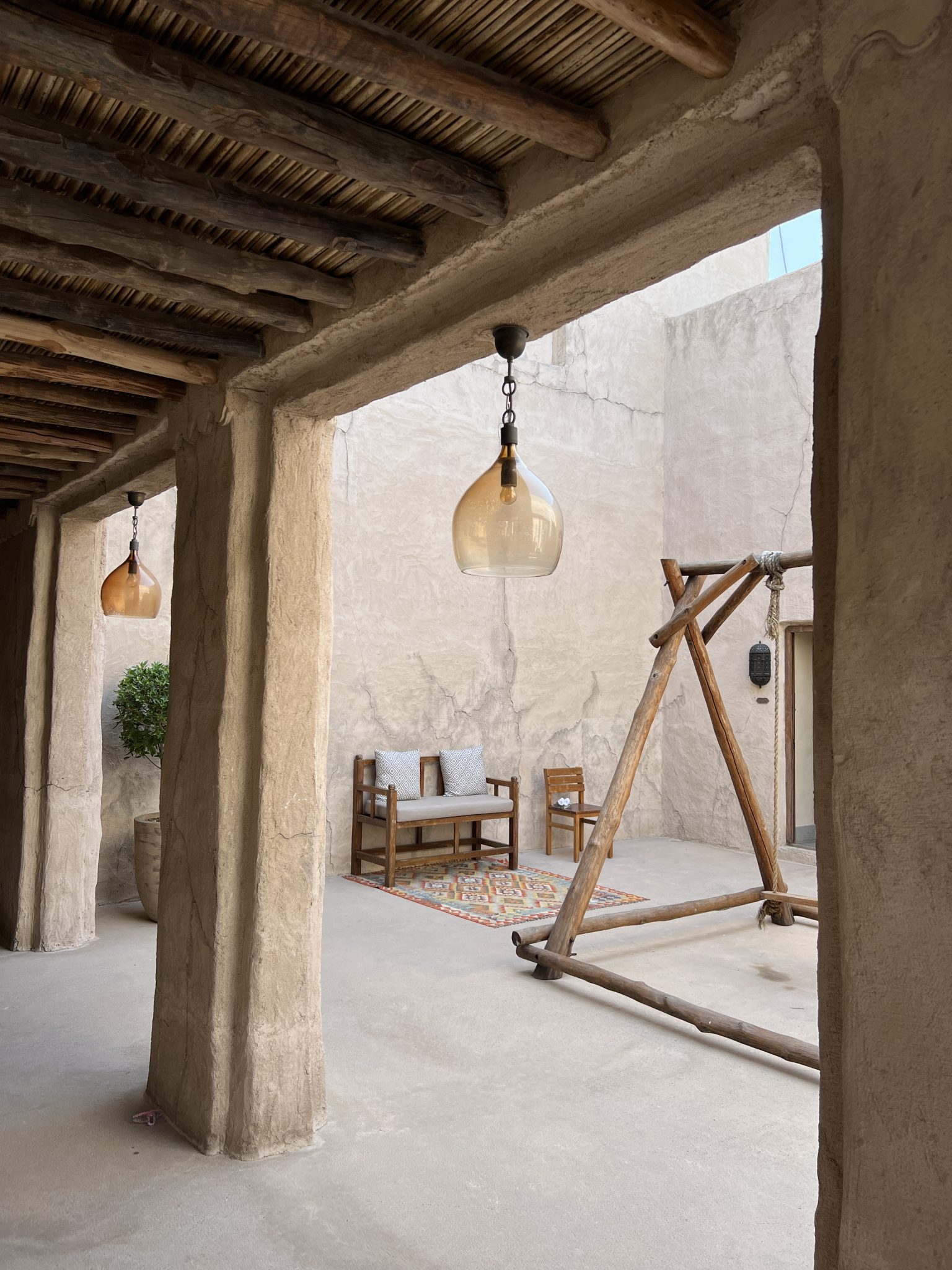 Two glass pendant lights hang from the ceiling. The courtyard features wooden outdoor furniture, including a bench with two cushions and a small chair.